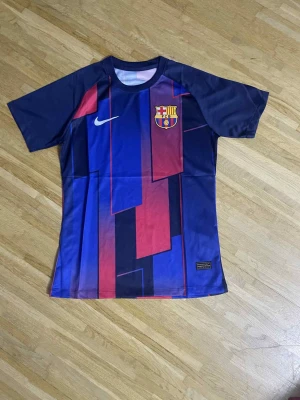 Barça Nike tröja med grafiskt mönster - Säljer en FC Barcelona tröja från Nike med ett fett grafiskt mönster i blått och rött. Tröjan har klubbmärket på bröstet och Nike-loggan. Materialet är polyester och designen är kortärmad med rund hals. Perfekt för dig som älskar fotboll och vill sticka ut.