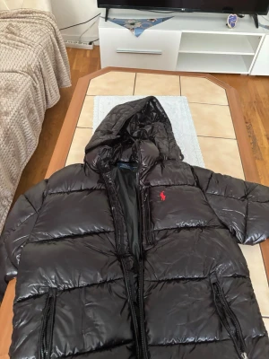 Svart pufferjacka från Polo Ralph Lauren - Svart pufferjacka med glansig finish från Polo Ralph Lauren. Jackan har huva, två sidofickor med dragkedja och det klassiska röda Polo-logot på bröstet. Perfekt för kalla dagar och har en riktigt schysst oversized look.