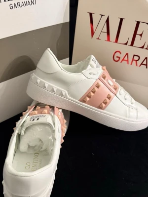 Valentino Rockstud sneakers vit/rosa - Säljer ett par Valentino Rockstud sneakers i vitt skinn med ljusrosa band och ikoniska nitar. Skorna har rund tå, platt sula och snörning framtill. Märkeslogga på plösen och insidan. Perfekt för dig som gillar lyxiga detaljer och streetstyle.