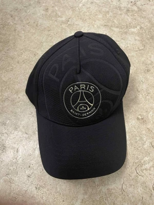 Svart Paris Saint-Germain keps - Snygg svart keps från Paris Saint-Germain med broderad logga framtill och texten 'C'EST PARIS' på baksidan. Justerbar rem för perfekt passform och detaljer med klubbnamnet på insidan. Perfekt för dig som gillar fotboll och vill ha en cool accessoar.