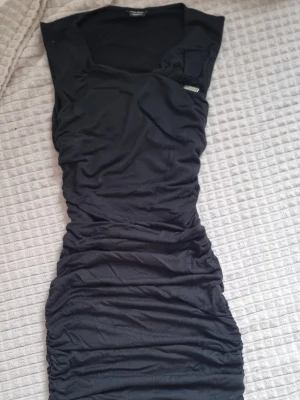 Svart kortklänning från Miss Sixty - Säljer en svart kortklänning från Miss Sixty i storlek XS. Klänningen har snygga rynkade detaljer längs sidorna och en v-ringad hals. Materialet är mjukt och stretchigt, vilket gör den bekväm att bära. Perfekt för dig som gillar en slim passform och stilren look.