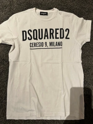 Vit DSQUARED2 t-shirt med tryck - Snygg vit t-shirt från DSQUARED2 med svart tryck framtill där det står 'DSQUARED2 CERESIO 9, MILANO'. Klassisk rund halsringning och korta ärmar. Tillverkad i mjuk bomull som är skön att ha på sig. Perfekt för dig som gillar stilrena och trendiga plagg.