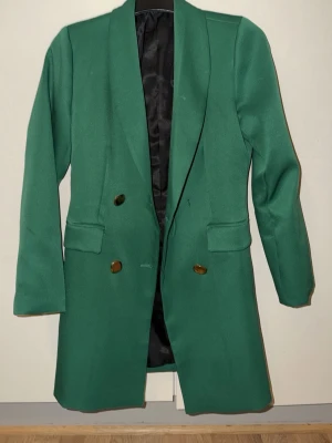 Grön kavaj med knappar - Snygg grön kavaj med klassisk passform och långa ärmar. Kavajen har två fickor framtill och dekorativa bruna knappar. Insidan är fodrad i svart tyg och modellen är lite längre, vilket ger en cool och stilren look.