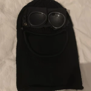 Svart stickad mössa med glasögon - Cool svart stickad mössa från C.P. Company med inbyggda glasögon. Perfekt för att hålla sig varm och se stilren ut. Mössan har en unik design med glasögon som kan fällas ner över ansiktet.