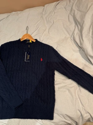 Mörkblå stickad tröja Polo Ralph Lauren - Kabelstickad mörkblå tröja från Polo Ralph Lauren med klassisk röd logga broderad på bröstet. Tröjan har rund halsringning, långa ärmar och ribbade muddar. Perfekt för dig som vill ha en tidlös och snygg look.