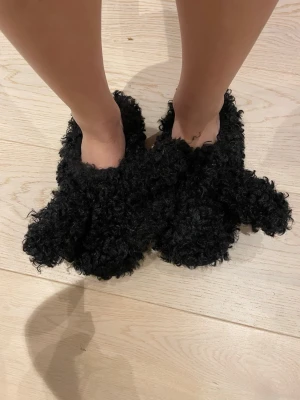 Svarta fluffiga tofflor med söta detaljer  - Supermysiga svarta tofflor med fluffigt pälsmaterial och en söt design . Perfekta för att hålla fötterna varma hemma, med en mjuk och skön känsla. Tofflorna har rund tå och är gjorda i syntetiskt pälsmaterial som ger en cool look.