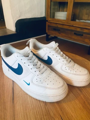 Nike Air force 1 royal blue - Nike Air Force 1 sneakers i vitt med blå swoosh och detaljer. Klassisk låg modell med snörning och perforerad tå för extra ventilation. Ovandel i skinn och textil, platt sula och rund tå. Perfekt för dig som gillar stilrena och sportiga sneakers.