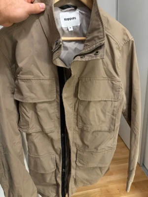 Beige overshirt från Kappahl -  Kappahl i storlek M. Jackan har fyra stora fickor framtill, dragkedja och knappar vid halsen. 