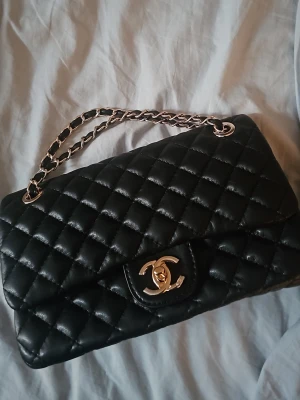 Svart quiltad handväska  - Säljer en ikonisk svart handväska  med quiltad design och guldfärgad kedja. Väskan är tillverkad i mjukt skinn och har det klassiska CC-spännet framtill. Insidan har dragkedja och präglad logga. Perfekt för dig som vill ha en lyxig och tidlös accessoar.
