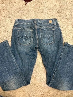 Blå bootcut jeans från Guess, storlek 28 - Säljer ett par klassiska blå jeans från Guess i bootcut-modell. Jeansen har fem fickor, snygga kontrastsömmar och broderade detaljer på bakfickorna. Materialet är slitstarkt denim i bomull och passformen är normal med låg midja.
