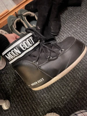 Svarta Moon Boots med snörning - Säljer ett par svarta Moon Boots med vit logga och snörning framtill. Skorna har en rund tå och platt sula, perfekt för kalla vinterdagar. Materialet är syntet och de har en klassisk moon boot-design med högt skaft.