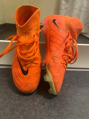 Nike Phantom Luna orange fotbollsskor - Säljer ett par Nike Phantom Luna fotbollsskor i stark orange färg med svarta Nike-loggor. Skorna har snörning, hög strumpkrage och en platt sula med dobbar för gräsplan. Ovandelen är stickad och syntet, designad för bra grepp och kontroll på planen.