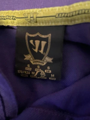 Lila Liverpool pikétröja från Warrior - Säljer en lila pikétröja från Warrior med Liverpool FC-logga och Union Jack-flagga på bröstet. Tröjan har vit kortärmad kontrast på ärmarna, klassisk krage och knappar framtill. Perfekt för dig som gillar fotboll och vill sticka ut med färg.