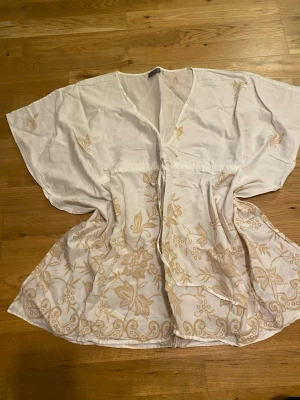 Vit blus med broderade blommor - Säljer en luftig vit blus med v-ringning och korta, vida ärmar. Blusen har ljusbeige broderade blommor och fjärilar samt en knytning i midjan. Perfekt för varma dagar och har en bohemisk vibe.
