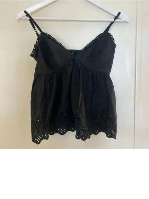 Bik bok topp - Säljer en svart linnetopp med tunna axelband och broderade spetsdetaljer längs kanten. Toppen har en luftig passform och vackert hålmönster nertill. Perfekt för varma dagar och enkel att matcha med jeans eller kjol.