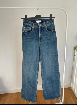 Blå wide jeans från H&M, strl 34 - Säljer ett par klassiska blå jeans från H&M i storlek 34. Modellen har hög midja och raka, vida ben. Jeansen har fem fickor, bälteshällor och stängs med knapp och dragkedja. Materialet är tålig denim i bomull.