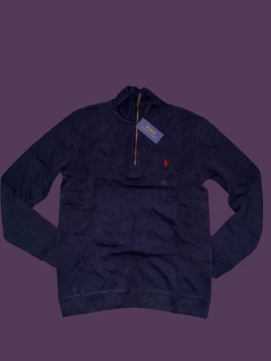 Marinblå half zip tröja Polo Ralph Lauren - Snygg marinblå tröja från Polo Ralph Lauren med half zip och ståkrage. Tröjan har ett rött broderat logomärke på bröstet och är tillverkad i mjuk bomull. Långärmad modell med normal passform, perfekt för chill höst- och vinterdagar.