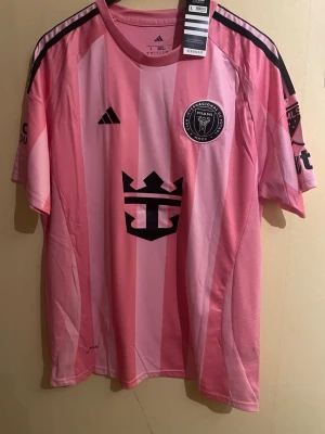 Inter Miami - Messi #10 - Set - Inter Miami-tröja i ikonisk rosa design, förknippad med Lionel Messi. Tröjan är i storlek L och har Adidas-logga, Inter Miami-emblem samt sponsortryck framtill. Skickas i mycket fint skick och passar perfekt för både match, vardag eller samling. Ett måste för Messi-fans eller MLS-intresserade. 