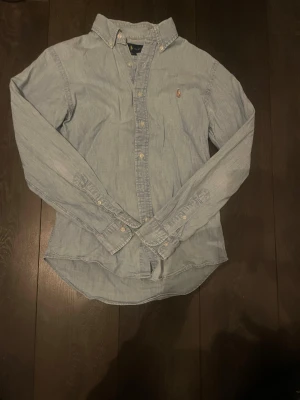 Ljusblå skjorta från Ralph Lauren - Säljer en ljusblå slim fit skjorta från Ralph Lauren med klassisk button-down krage och broderad logga på bröstet. Skjortan är långärmad och har knappar hela vägen framtill. Perfekt för en clean och stilren look.