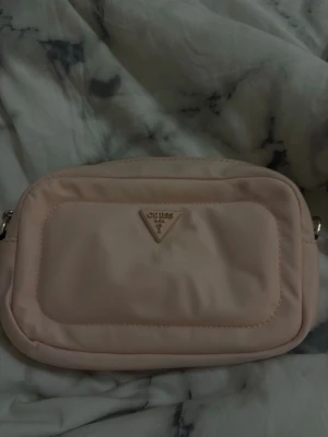 Ljusrosa axelväska från Guess - Säljer en ljusrosa axelväska från Guess med guldfärgad kedja och detaljer. Väskan har ett stort fack, innerficka med dragkedja och ytterficka med dragkedja. Materialet är mjuk polyester och väskan har Guess-logga i metall framtill. Fick väskan ( present ) från Australien 