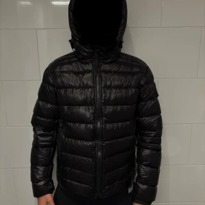 Canada Goose crofton puffer black label - Svart pufferjacka från Canada Goose, modell Crofton Hoody. Jackan har huva med snörning, två sidofickor med dragkedja och ett emblem på ärmen. Materialet är glansig polyester och dunfyllning. Snygga reflexdetaljer på huvan och stilren design.