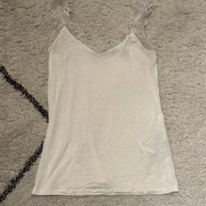 Vit linnetopp med spetsband - Säljer en vit linnetopp med v-ringad hals och breda axelband i spets. Toppen är ärmlös och har en enkel, stilren design som passar perfekt till sommaren. Materialet känns lätt och luftigt, och spetsdetaljen ger en extra touch.