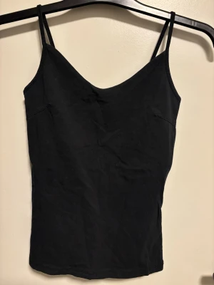 Svart linnetopp med smala axelband - En stilren svart linnetopp med smala justerbara axelband och v-ringad hals. Toppen är ärmlös och har en enkel, tight passform som sitter snyggt mot kroppen. Materialet känns mjukt och lätt, perfekt för varma dagar eller som lager under en skjorta.