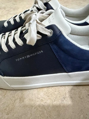 Tommy Hilfiger sneakers i marinblått - Snygga sneakers från Tommy Hilfiger i marinblått och vitt med rund tå och platt sula. Skorna har vita snören, vit häl och detaljer med logga på sidan och bak. Materialet är en mix av skinn och syntet, vilket ger en fräsch och stilren look.Ny kostar 1399kr,Den använd en gång 
