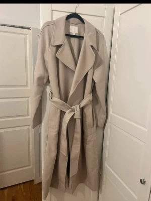 Beige trenchcoat från H&M med bälte - Oanvänd utan prislap. Snygg beige trenchcoat från H&M i storlek S. Jackan har klassisk krage, långa ärmar och ett matchande bälte i midjan. Fickor på sidorna och stilren design gör den enkel att styla. Perfekt för dig som gillar minimalistiskt och cleant.