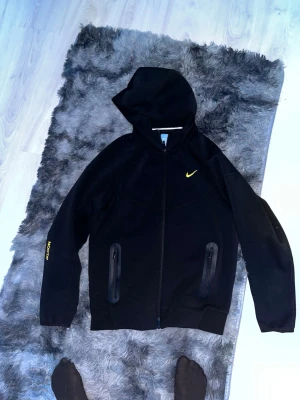 Nice nocta tech fleece  - Säljer en svart vindjacka från Nike med gul logga på bröstet. Jackan har huva, hel dragkedja framtill samt två fickor med dragkedja. En stilren och bekväm jacka med sportig look – perfekt för både vardag och träning. I mycket gott skick.