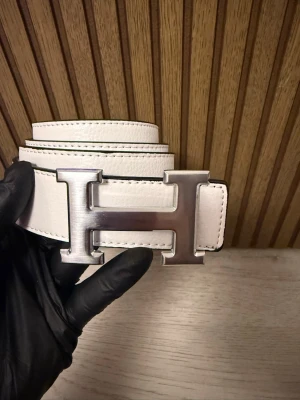 Vit skinnbälte med H-spänne - Säljer ett stilrent vitt bälte med ett stort H-format metallspänne i silver. Bältet är gjort i äkta skinn med tydliga sömmar och har en svart insida. Perfekt för att lyfta din outfit med en lyxig touch.