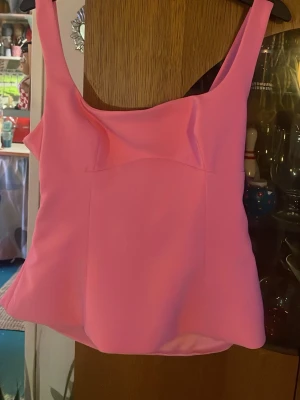 Rosa linnetopp med rosett och öppen rygg - Säljer en ljusrosa linnetopp med breda axelband och en snygg rosett bak. Toppen har en öppen rygg med ett hål under rosetten och är tillverkad i ett slätt syntetmaterial. Perfekt för dig som gillar färgglada och trendiga plagg. Snygg både till vardag och fest. 