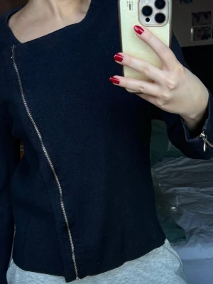 Svart stickad kofta med sned dragkedja - Säljer en svart stickad kofta med unik snedställd dragkedja framtill. Koftan har långärmade ärmar och en cool asymmetrisk halsringning. Perfekt för dig som gillar stilrena detaljer och vill ha något som sticker ut lite extra. Den är storlek XL men passar mig som vanligtvis är S.