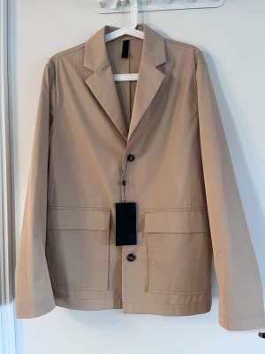 J.Lindeberg Cotton Blazer - Beige blazer från J.Lindeberg i lätt bomull. Klassisk krage, två stora fickor framtill och knäppning med knappar. Storlek S med boxig passform. Perfekt för dig som gillar stilrena och enkla plagg med modern touch. Passar dig som vill ha en clean look. Helt ny, med prislapp! Säljs på flera sidor, först till kvarn. 