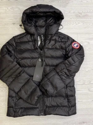 Canada goose crofton jacket  - Säljer en svart, quiltad dunjacka från Canada Goose med huva och dragkedja framtill. Jackan har ett snyggt märke på ärmen och är perfekt för kalla dagar. Den har en modern, puffad look och är riktigt skön att bära. Passformen är normal och jackan är långärmad.