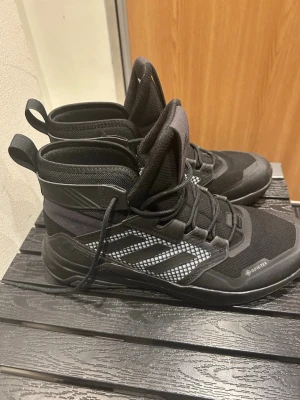 Svarta Adidas Terrex Gore-Tex kängor - Säljer ett par svarta Adidas Terrex kängor med Gore-Tex membran. Skorna har snörning, meshpaneler med rutigt mönster och robust platt sula. Perfekta för utomhusaktiviteter och håller fötterna torra. Märket syns tydligt på plösen och innersulan.