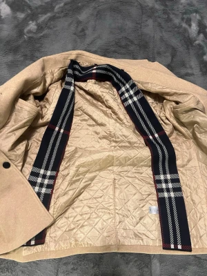 Beige kappa med rutigt foder - Snygg beige ull kappa från  med klassiskt rutigt foder i svart, vitt och rött. Jackan har rak passform, svarta knappar framtill och hög krage. Utsidan är i ullmix och insidan quiltad för extra värme. Perfekt för dig som vill ha en stilren och tidlös look. Ingen tecken av användning. Inte i orginal förpackning 