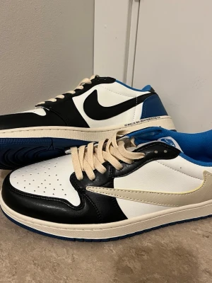 Nike Air Jordan 1 Low Travis Scott x Fragment - Nike Air Jordan 1 Low Travis Scott x Fragment med svart, vit och blå färg. Skorna har omvänd vit swoosh på utsidan, klassisk svart swoosh på insidan, blå yttersula och detaljer som Cactus Jack och Fragment-logga på hälen. Tillverkade i skinn med beige snörning och perforerad tå.
