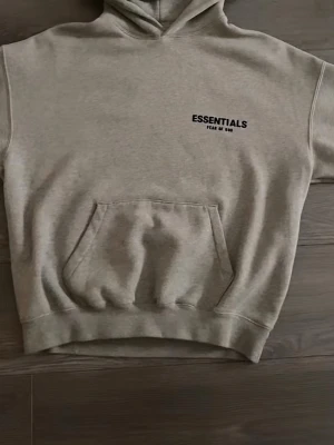 Essentials Hoodie  - Snygg Essentials (Fear of God) hoodie i beige. Minimalistisk design med Essentials-logga på bröstet. Mjuk och bekväm passform, perfekt för vardag och streetwear.