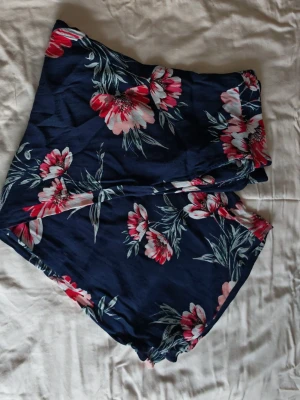 Blommiga byxshorts från Lindex, storlek L - Säljer ett par mörkblå byxshorts från Lindex i storlek L. De har ett färgglatt blommigt mönster i rosa, vitt och grönt samt elastisk midja för extra komfort. Innermått 67 cm. Tillverkade i 100% viskos vilket gör dem lätta och luftiga. Perfekta för varma dagar!