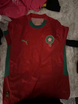 Marocko fotbolls tröja  - Bilderna visar en röd och grön fotbollströja från Puma med Marockos landslagsemblem, men inga skor syns på bilderna.