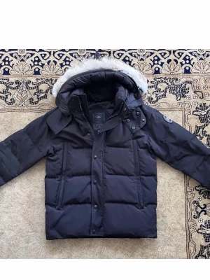 Canada Goose wyndham - Säljer en svart pufferjacka från Canada Goose med avtagbar huva och pälskant. Jackan har klassisk patch på ärmen, dragkedja och knappar framtill samt två stora fickor. Perfekt för kalla vinterdagar och riktigt snygg streetstil.