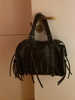 Svart axelväska med fransar - Cool svart axelväska i skinn med snygga fransdetaljer och silverfärgade metalldelar. Väskan har både handtag och justerbar axelrem, samt en praktisk dragkedja framtill. Perfekt för dig som gillar en edgy stil och vill ha plats för allt du behöver.