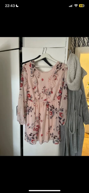 Ljusrosa blommig kortklänning med volang - Säljer en ljusrosa kortklänning med blommigt mönster i rött, blått och beige. Klänningen har v-ringning, volangdetaljer framtill och långa, transparenta ärmar. Materialet känns lätt och luftigt, perfekt för varmare dagar.