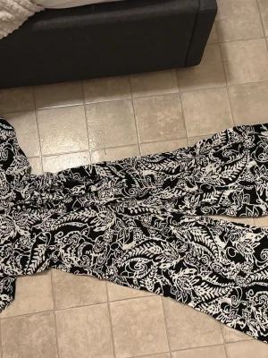 Svarta wide byxdress från Amisu - Säljer en svart och vit byxdress från Amisu i storlek 34. Byxdressen har ett blommigt och paisley-inspirerat mönster, vida ben och v-ringad hals. Materialet är mjukt och luftigt, perfekt för varmare dagar. Supersnygg och skön att bära!