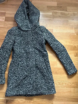 Gråmelerad ullkappa med huva från ONLY - Säljer en gråmelerad lång kappa från ONLY i storlek XS. Jackan har en stor och mysig huva, långärmade ärmar och är tillverkad i ullblandning. 