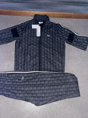 Mörkgrå mönstrad Lacoste tracksuit - Säljer en mörkgrå tracksuit från Lacoste med geometriskt mönster och vita detaljer längs ärmar och ben. Setet har dragkedja på jackan och elastisk midja på byxorna. Klassisk Lacoste-logga på både jacka och byxa. Perfekt för en sportig och stilren look.