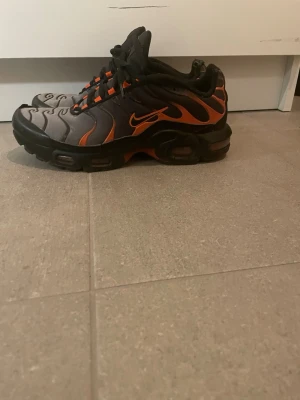 Nike Air Max Plus Tn svart och orange - Nike Air Max Plus Tn sneakers i svart och orange med coola vågiga detaljer på ovansidan och Tn Air-logga bak. Skorna har snörning, platt sula och rund tå. Materialet är syntet och mesh, vilket gör dem både lätta och bekväma. Perfekt för dig som gillar streetwear och sportig stil.
