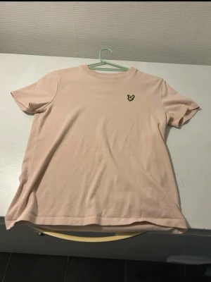 Ljusrosa t-shirt från Lyle & Scott - Säljer en ljusrosa t-shirt från Lyle & Scott med klassisk rund hals och korta ärmar. T-shirten har en liten broderad gul logga på bröstet och är gjord i mjuk bomull. Perfekt för dig som gillar stilrena och enkla plagg med en touch av märke.
