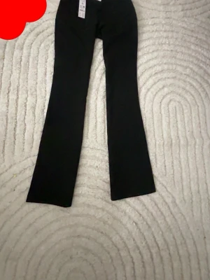 Svarta bootcut-byxor från Gina Tricot - Svarta bootcut-byxor från Gina Tricot i storlek 164. Snyggt lågmidjade med klassisk knapp och dragkedja framtill. Byxorna har en stilren look och är gjorda i ett mjukt material som sitter skönt. Perfekta för dig som gillar trendiga och tidlösa plagg.